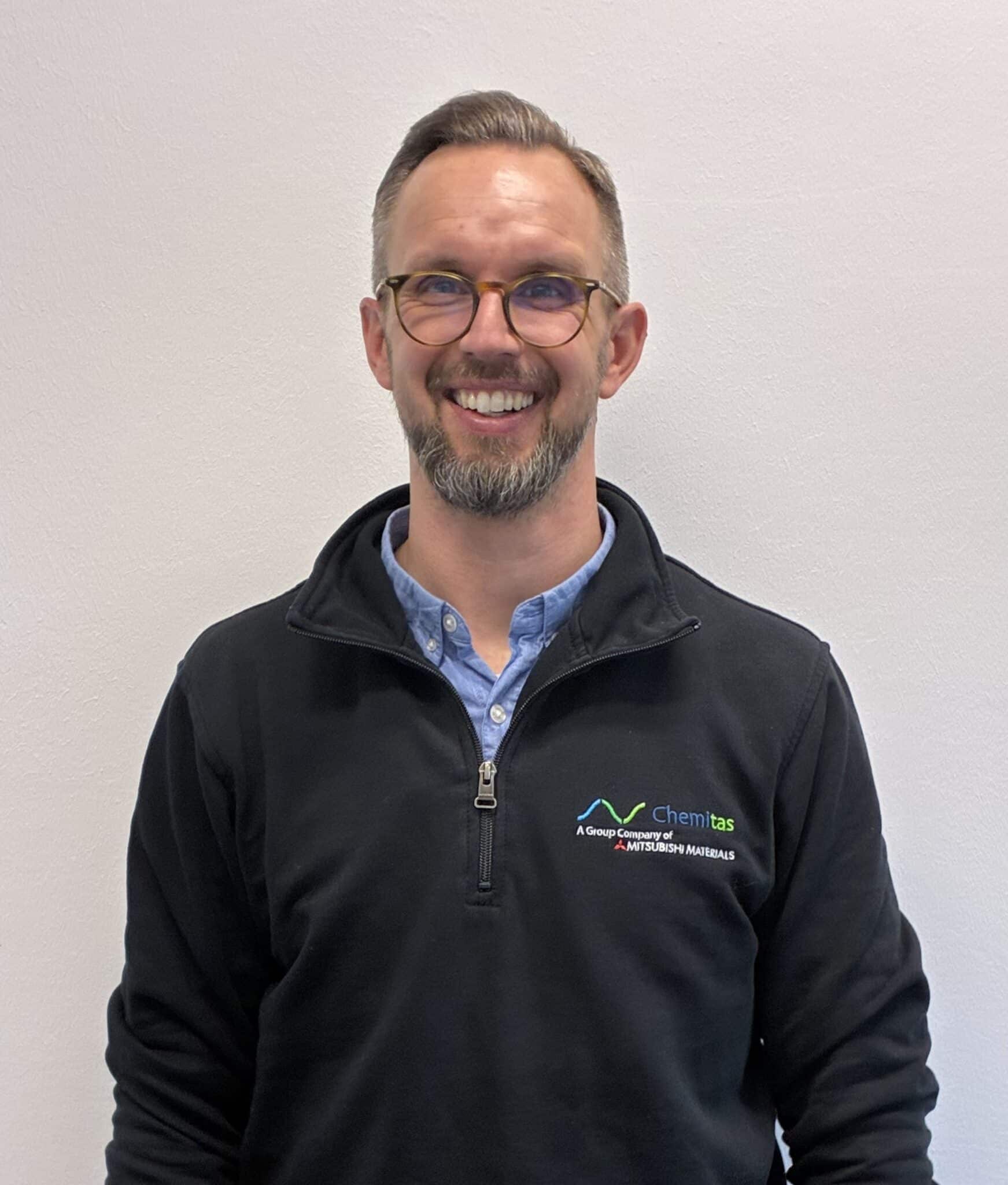 Ein lächelnder Mann mit kurzen Haaren, Brille und Bart steht vor einem schlichten hellen Hintergrund. Er trägt einen schwarzen Pullover mit Viertelreißverschluss und dem Firmenlogo von Chemitas über einem hellblauen Hemd.